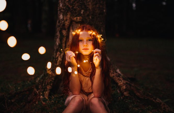 Mädchen, mit traurigem Blick zum Betrachter, eine Lichterkette die ins Bild führt, um ihren Kopf führt und sie in in ihren Händen hält, im Hintergrund ein Baum, an dem sie sitzt